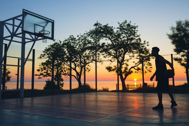 Черный человек занимается спортом, играет в баскетбол на восходе солнца, силуэт
