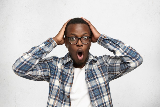 Free photo black man dressed casually, expressing shock and despair, touching his head with both hands, screaming with mouth wide opened and eyes popped