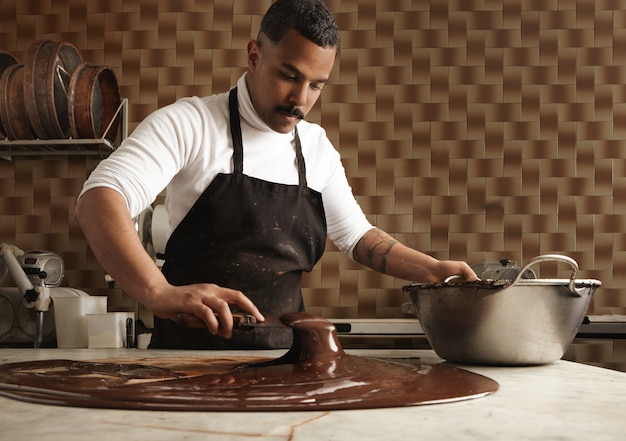 Black man professional chief collects tasty melted chocolate from vintage marble table to big steel pot in his artisan rustic kitchen with industrial retro machines.