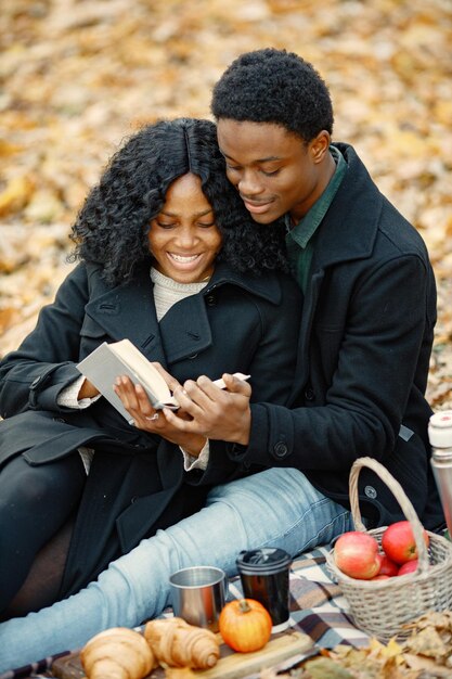 黒人男性と女性が抱き締めて本を読んでいます。秋の公園で毛布の上に座っているロマンチックなカップル。黒のコートを着た男と女。