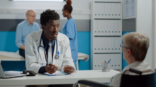 Black medic talking to disabled patient with handicap at desk in healthcare office. African american doctor helping elder woman with disability sitting in wheelchair. Old person at checkup