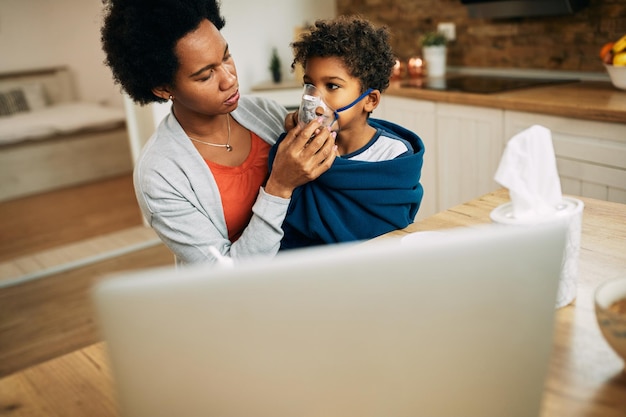 Black mother using nebulizer during inhaling therapy of her ill son at home