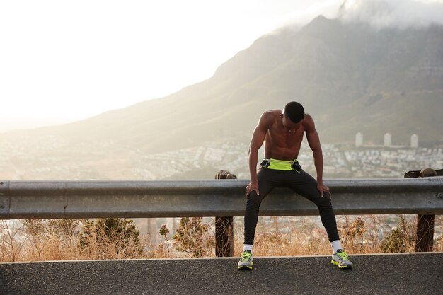 Black motivated sportsman feels exhausted after active fitness training, enjoys free lifestyle, rests after morning jog, keeps gaze down, has short hair, muscular athlete body, exercises outside