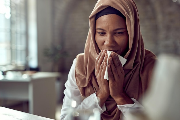 Черная мусульманская деловая женщина чихает в салфетку во время работы в офисе