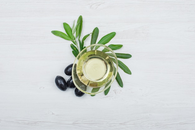 Black olives and olive oil in a glass can with olive leaves top view on white wood plank