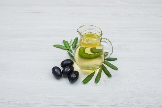 Black olives and olive oil in a glass jar with olive leaves side view on white wood plank