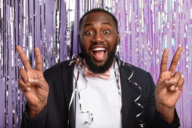 Black optimistic plump man with thick bristle, makes peace gesture, smiles positively, wears elegant suit, has serpentine on body, poses over purple wall with tinsel. Body language concept.