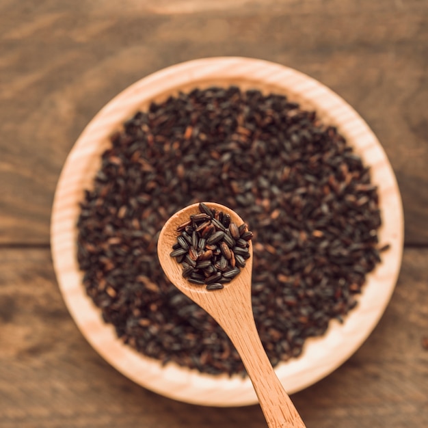 Black organic rice grain in spoon over the bowl on wooden backdrop