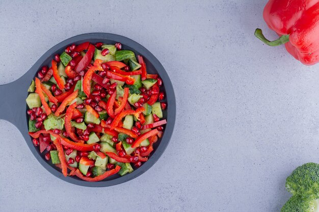 Черная сковорода овощного салата рядом с болгарским перцем и брокколи на мраморном фоне. Фото высокого качества