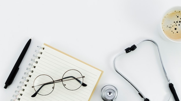 Black pen; eyeglasses on spiral notepad; stethoscope and coffee cup on white backdrop