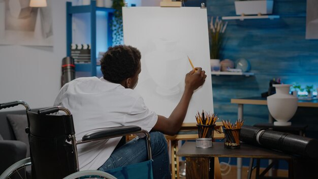Black person sitting in artwork space drawing vase using white canvas and pencil from table. Disabled african american artist in wheelchair creating design as artistic hobby