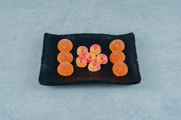 Черная тарелка, полная круглых апельсиновых желейных конфет в форме колец и апельсиновых желейных конфет с сахаром.