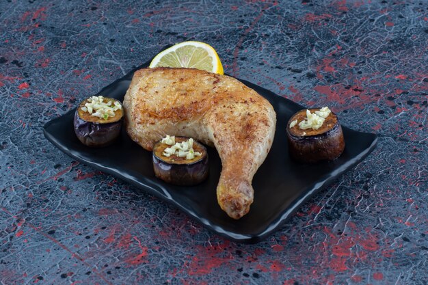 A black plate of grilled chicken leg with fried eggplant