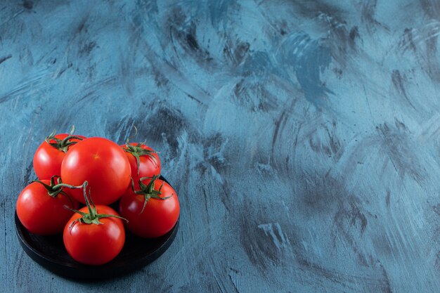 Black plate of red fresh tomatoes on blue background.