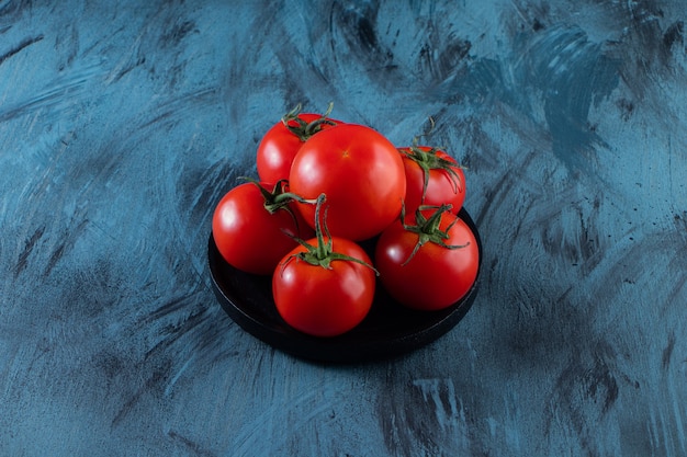 Black plate of red fresh tomatoes on blue surface.