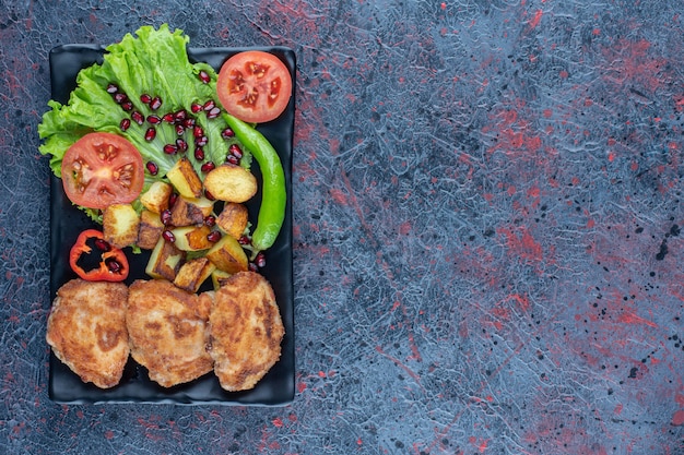 A black plate of vegetables and chicken cutlets . 