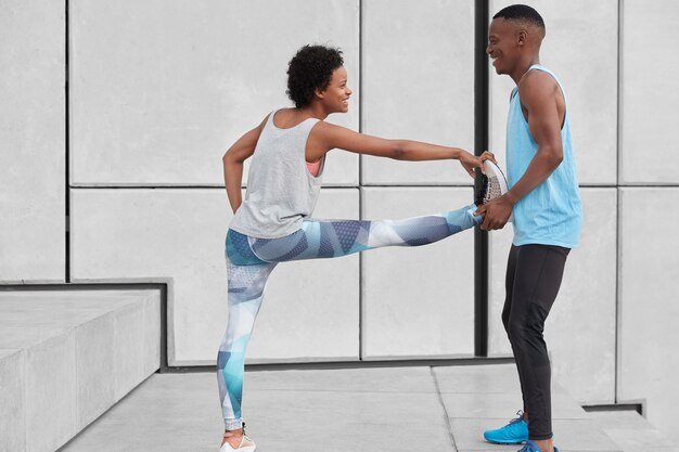 Black positive male instructor help female trainee to do exercises for flexibility, stand at stairs against white wall, have happy expressions, dressed in sportswear. People, sport and traning concept