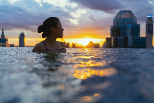 アジアの女性の黒いシルエットは、夏休みの休日に水をはねかける青い海の夕日の景色を望むインフィニティプールでリラックスできます。高層の空の景色都市のダウンタウン健康的な幸せのライフスタイル