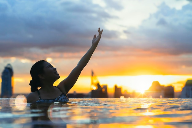 Siluetta nera della donna asiatica spruzzata d'acqua durante le vacanze estive rilassante nella piscina a sfioro con vista al tramonto sul mare blu con grattacieli del centro urbano del centro urbano sano stile di vita di felicità