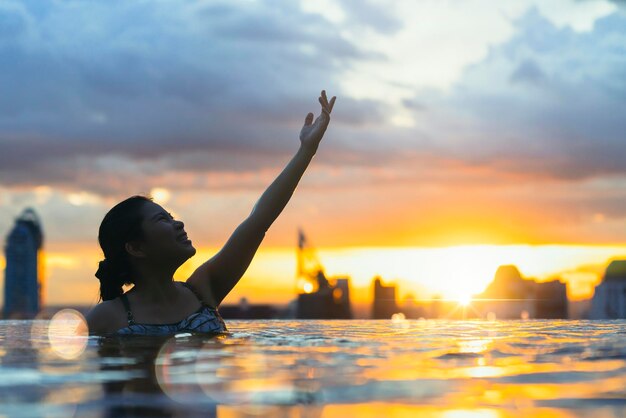 アジアの女性の黒いシルエットは、夏休みの休日に水をはねかける青い海の夕日の景色を望むインフィニティプールでリラックスできます。高層の空の景色都市のダウンタウン健康的な幸せのライフスタイル