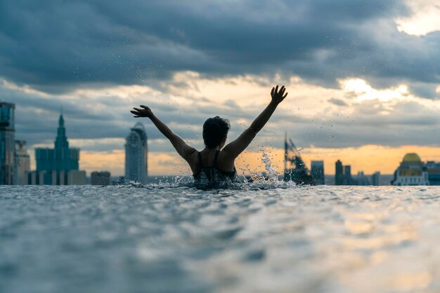 アジアの女性の黒いシルエットは、夏休みの休日に水をはねかける青い海の夕日の景色を望むインフィニティプールでリラックスできます。高層の空の景色都市のダウンタウン健康的な幸せのライフスタイル