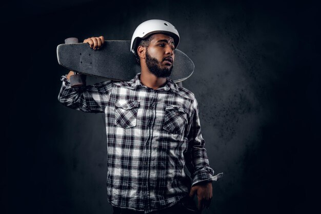 Black skater dressed in a fleece shirt holds long board over grey background in a studio.