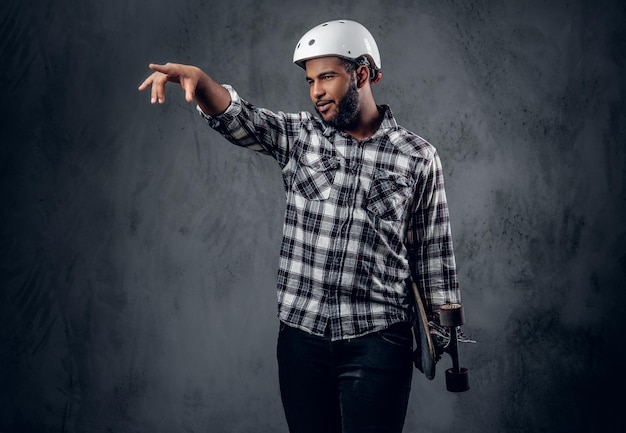 Black skater dressed in a fleece shirt holds long board over grey background in a studio.