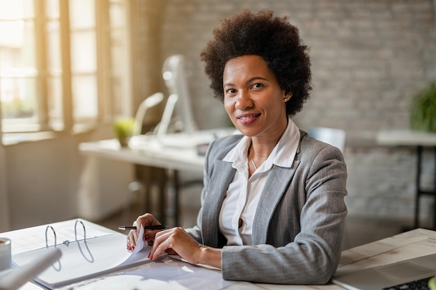 Черная улыбающаяся деловая женщина анализирует финансовые отчеты во время работы в офисе и смотрит в камеру