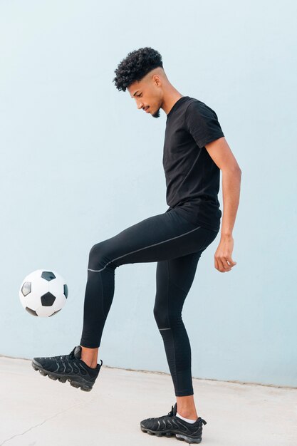 Black sportsman kicking football at blue wall background