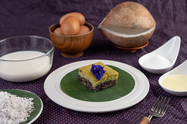 Black sticky rice and custard on a banana leaf in a white plate with butterfly pea flowers.