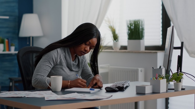 Black student writing education ideas on stickey notes sitting at desk table in living room