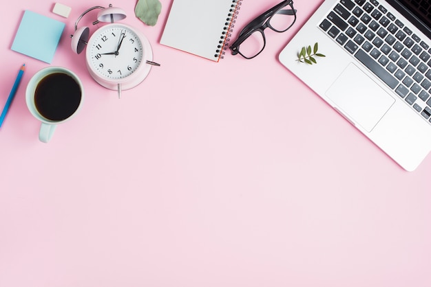 Black tea cup; alarm clock; spiral notepad; eyeglasses and laptop on pink backdrop