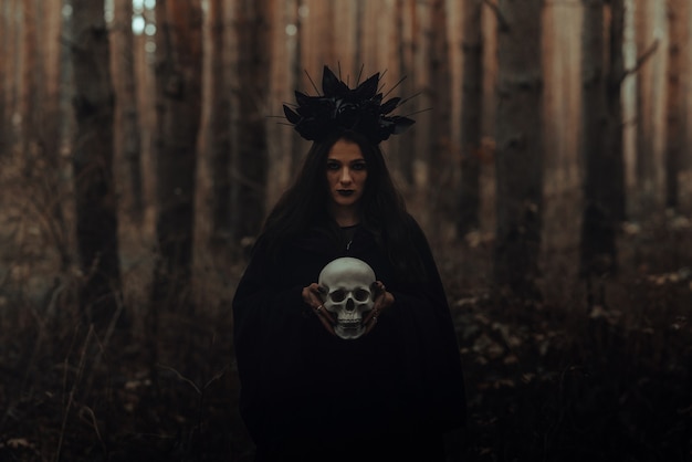 Black terrible witch holds the skull of a dead man in her hands in a dark forest