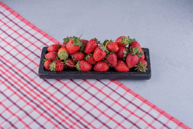 Black tray with a serving of fresh strawberries on tablecloth on marble background.