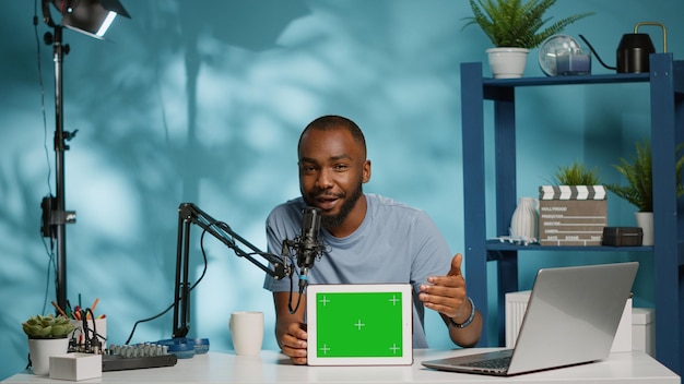 Black vlogger showing tablet with horizontal green screen on camera for podcast. African american man using chroma key with mockup template on digital gadget for vlog. Social media blogger