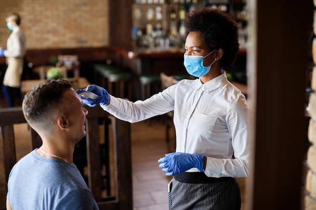 Free photo black waitress measuring temperature of a guest with infrared thermometer during coronavirus epidemic