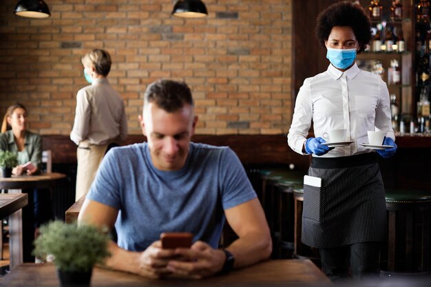 Black waitress wearing face mask and gloves while serving coffee in a cafe during coronavirus epidemic