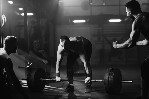 Black and white photo of determined athlete lifting barbell in a gym while his athletic friends are supporting him