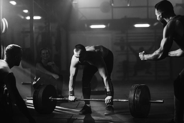 Black and white photo of determined athlete lifting barbell in a gym while his athletic friends are supporting him