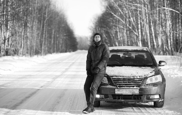 Black and white photo of winter rural road in forest in sunny day and man with car