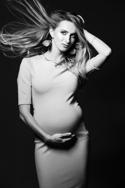 Black and white portrait of beautiful mom embracing stomach smiling looking away over shoulder Young happy mother posing at studio against dark background Pregnant fashion