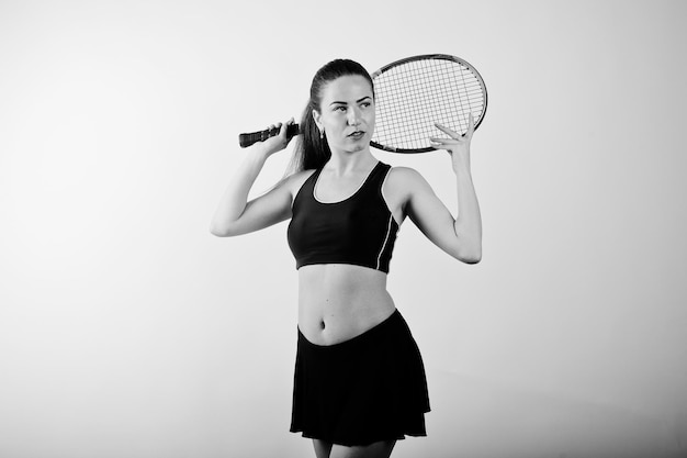 Black and white portrait of beautiful young woman player in sports clothes holding tennis racket while standing against white background