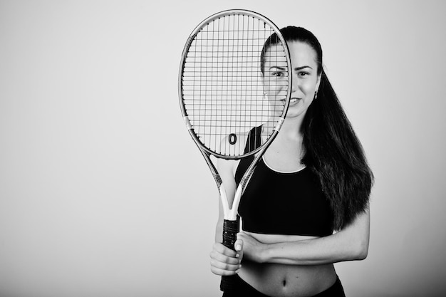 Black and white portrait of beautiful young woman player in sports clothes holding tennis racket while standing against white background