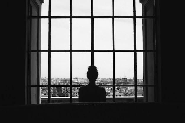 Black and white shot of a lonely female standing in front of the windows looking at the buildings