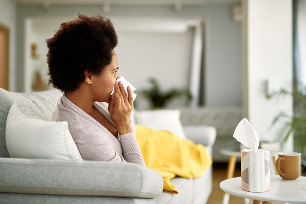 Black woman feeling sick and blowing her nose in a tissue while resting on in the living room