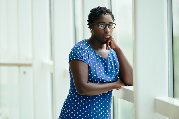 Black woman in glasses and polka-dot dress standing by window with head resting on hand