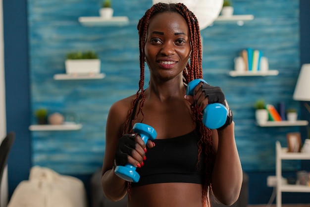 Black woman taking care of body working training arm muscles using dumbbells weights during workout. Positive joyful sportive strong fit athlete in home living room.