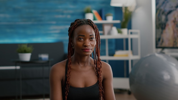 Black woman trainer sitting in lotus position on yoga mat on floor with closed eyes