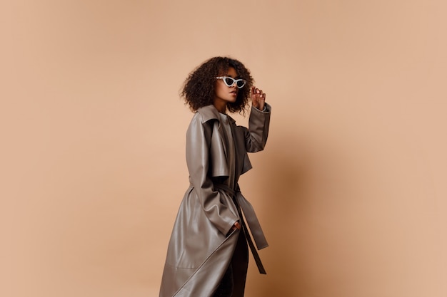 Free photo black woman in trendy grey leather jacket posing over beige background in studio. winter and autumn fashion look.