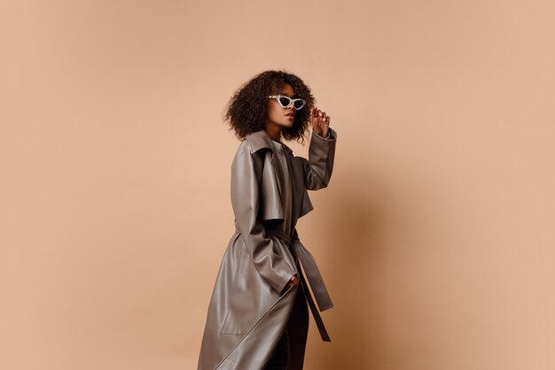 Black woman in trendy grey leather jacket posing over beige background in studio. Winter and autumn fashion look.